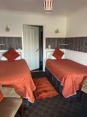 A hotel room with two beds, a wooden chair, a red rug, and a door leading to another room.
