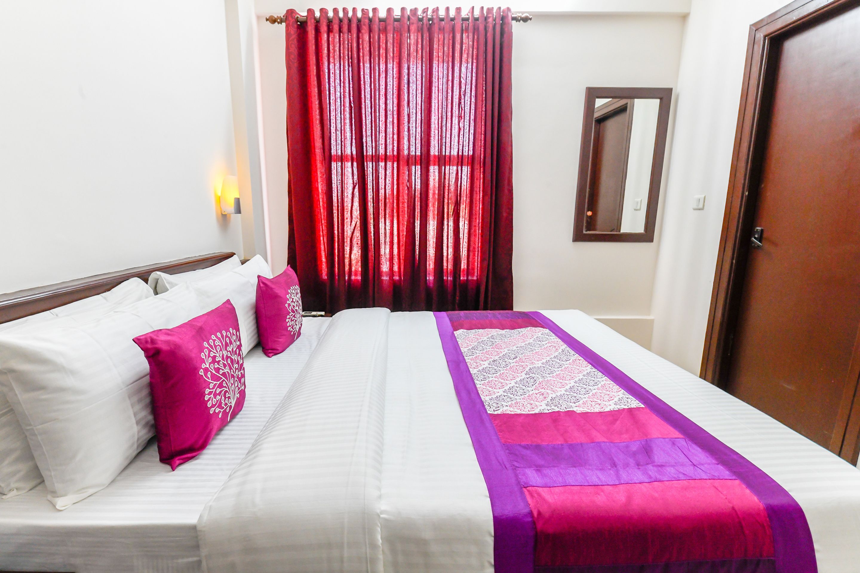 Een net opgemaakt bed met wit en roze beddengoed, een paars en roze quilt en een raam met rode gordijnen.