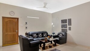 Una sala de estar con sofá, una mesa de centro, una puerta de madera, un ventilador de techo y obras de arte colgadas en la pared.
