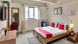 Habitación de hotel con cama, escritorio, silla, mesita de noche, lámpara, espejo, ventana con cortinas y ventilador de techo.