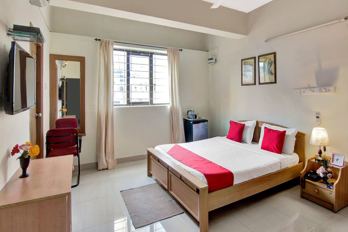 A hotel room with a bed, a desk, a chair, a nightstand, a lamp, a mirror, a window with curtains, and a ceiling fan.