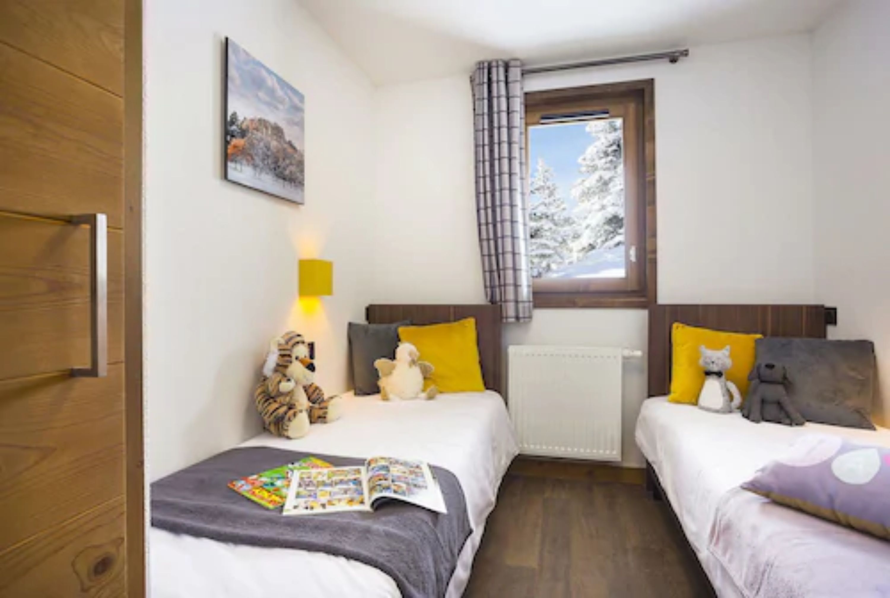 A bedroom with two beds, a window with a view of snow-covered trees, a radiator, and a painting on the wall.