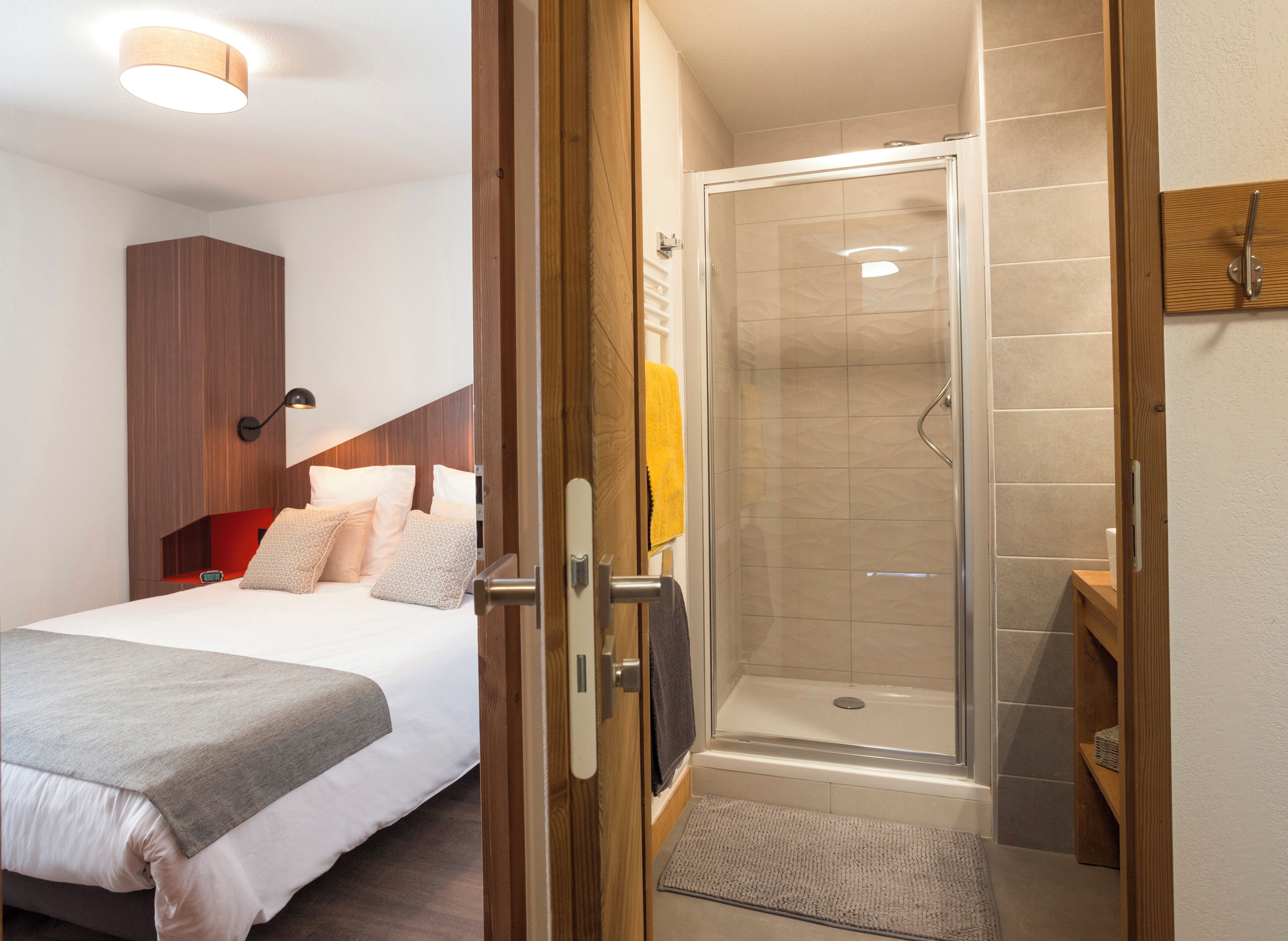 A hotel room with a bed, a wooden wardrobe, a shower area with a glass door, and a small wooden shelf.