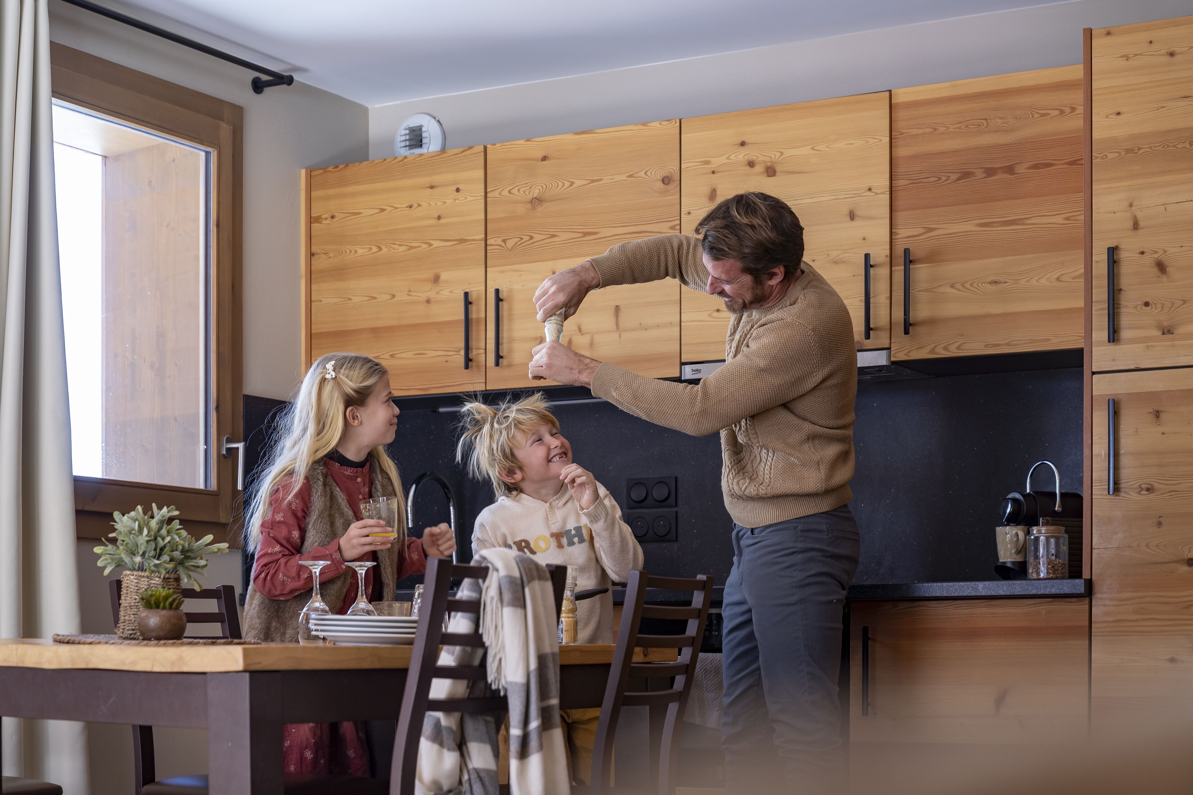 Ett modernt kök med träskåp, ett matbord med stolar och en person som häller upp en drink.