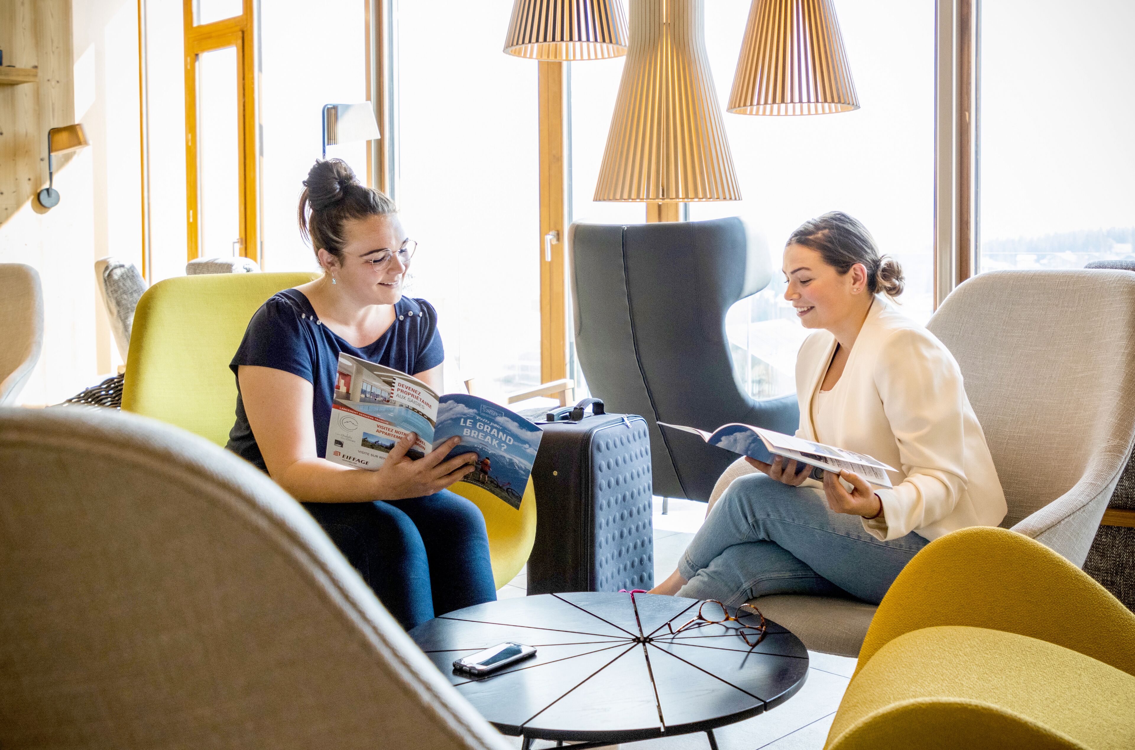 Two people sitting on chairs, reading a magazine. There is a table with a phone and a lamp between them.