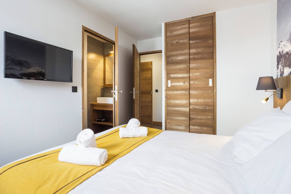 Een hotelkamer met een bed, een aan de muur bevestigde televisie, een houten garderobe en een badkamer die door een open deur zichtbaar is.