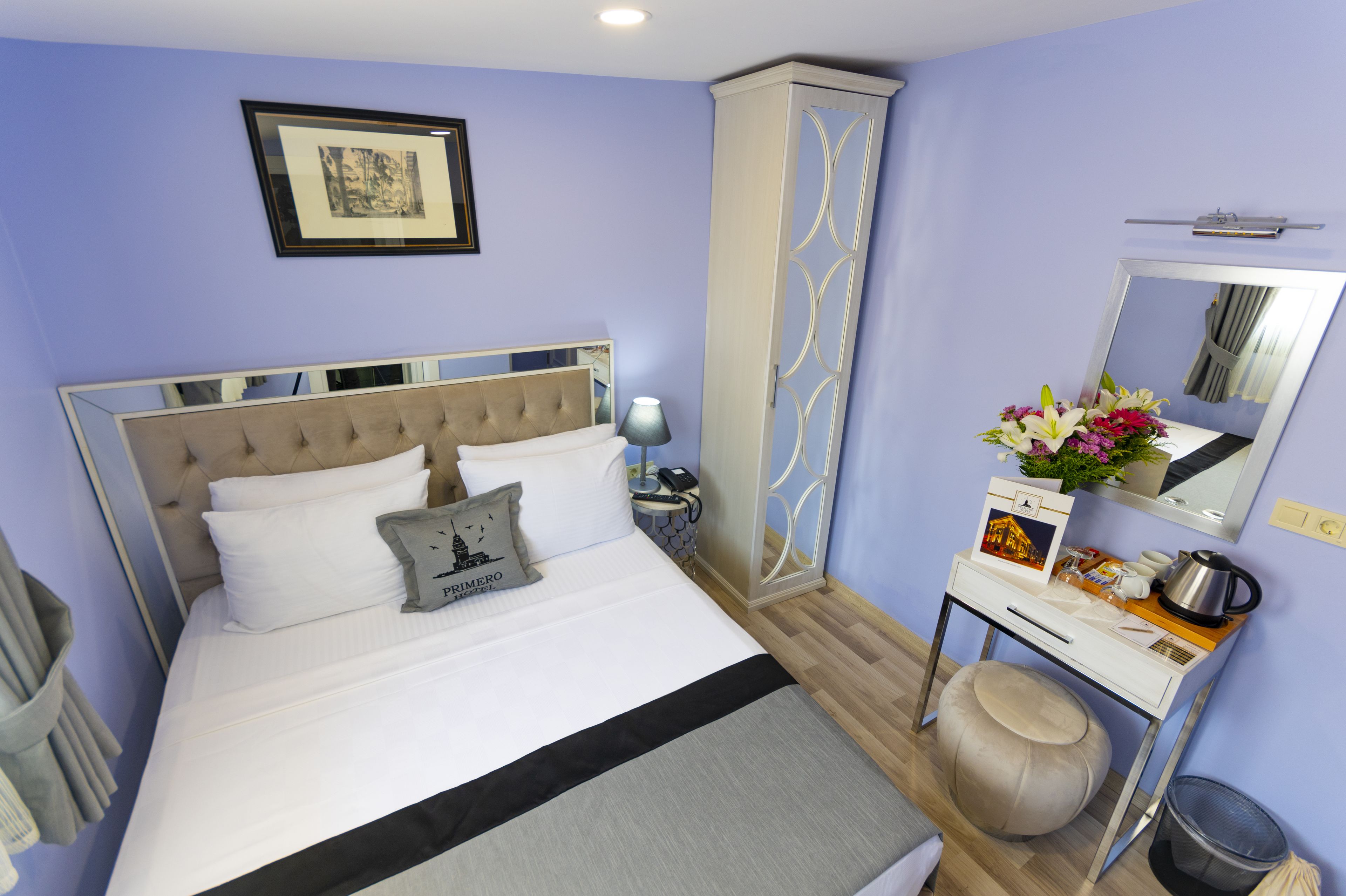 A neatly arranged hotel room with a bed, bedside table, lamp, wardrobe, mirror, and a small desk with a kettle and flowers.
