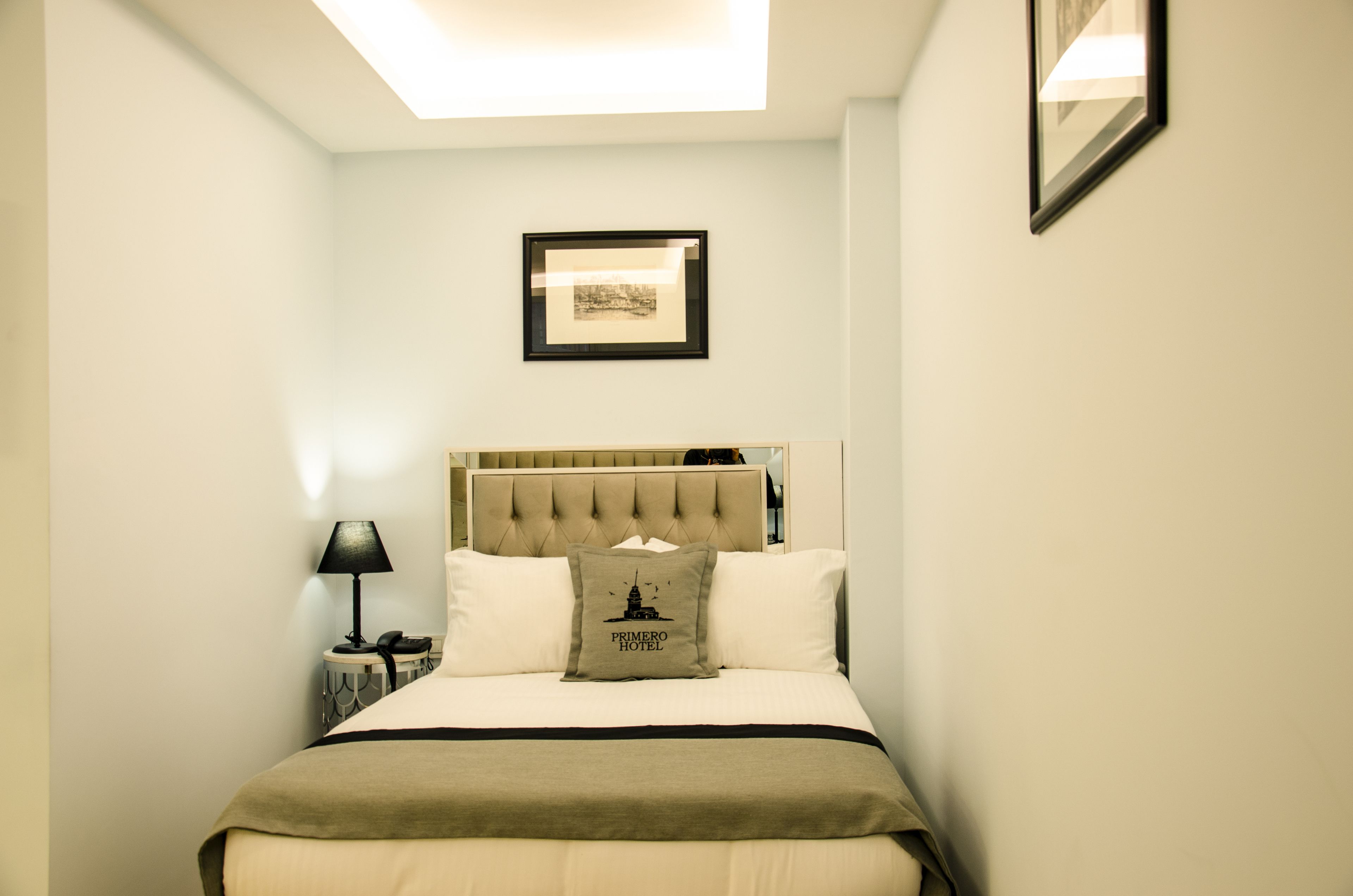 Quarto arrumado com cama, mesa de cabeceira com abajur, quadro na parede e travesseiro com o logotipo do hotel.