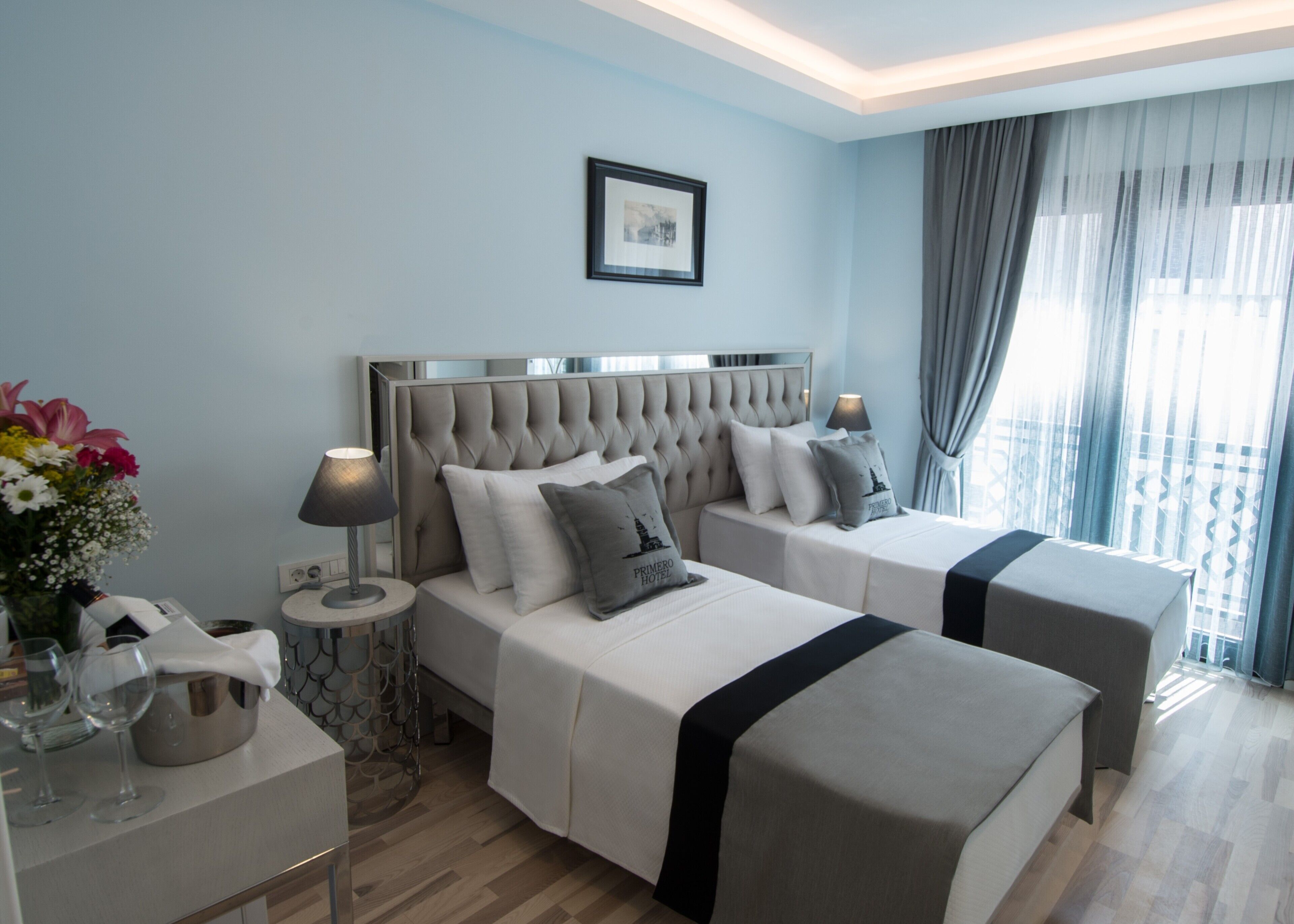 Habitación de hotel con dos camas, una mesita de noche con lámpara, un florero con flores y un cuadro en la pared.