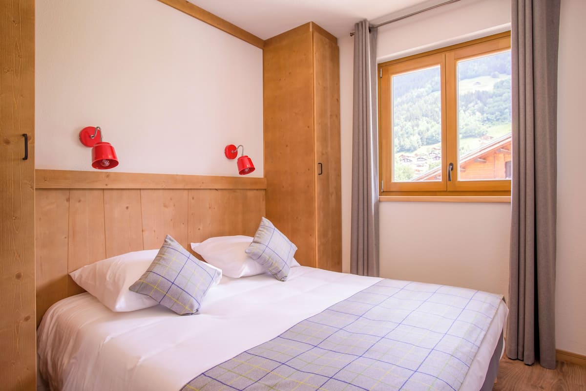 Een slaapkamer met een houten hoofdbord, een bed met witte beddengoed en geruite kussens, twee rode wandlampen en een raam met gordijnen.
