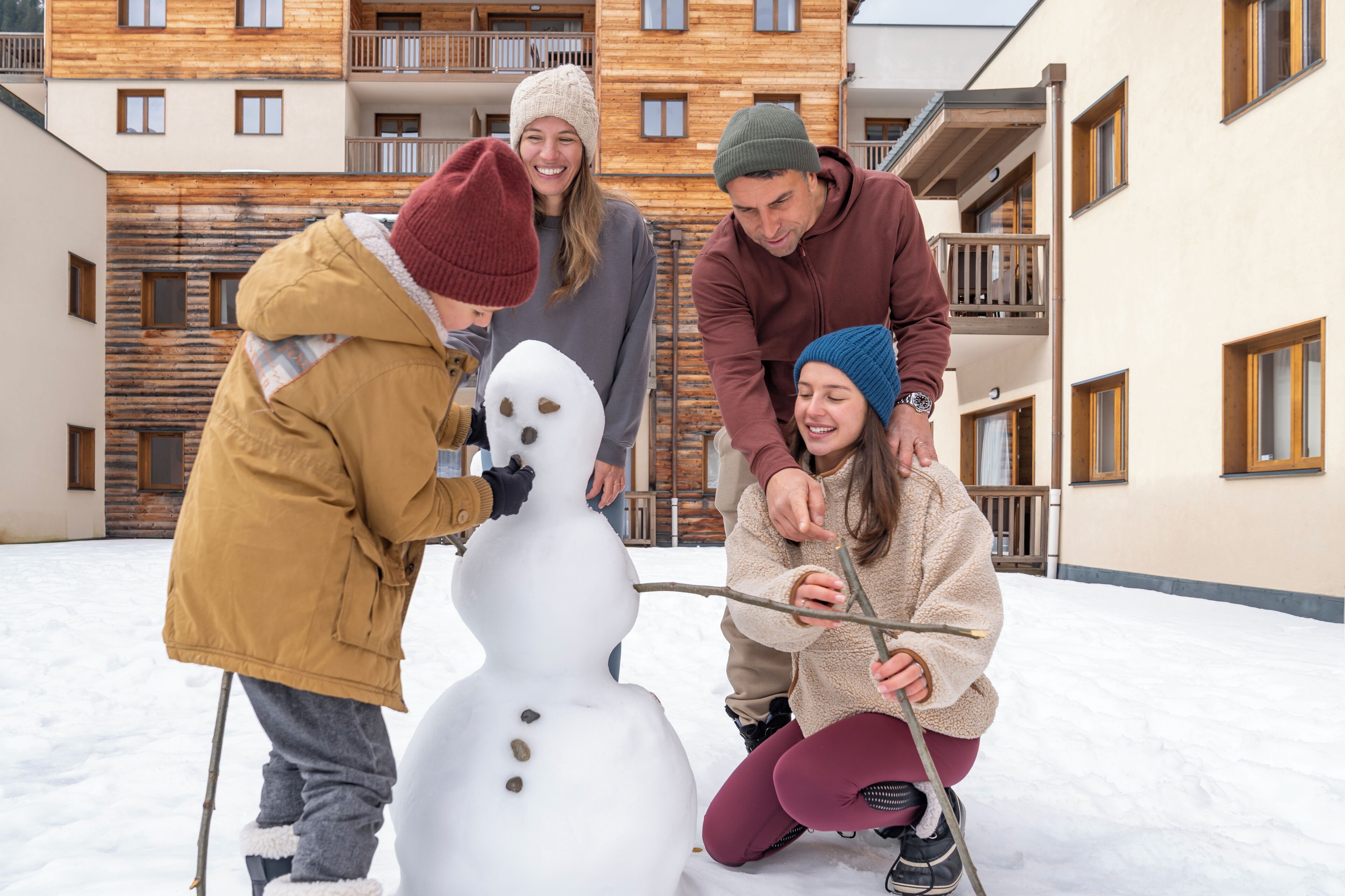 Cuatro personas construyen un muñeco de nieve en un espacio al aire libre, con edificios de madera al fondo.