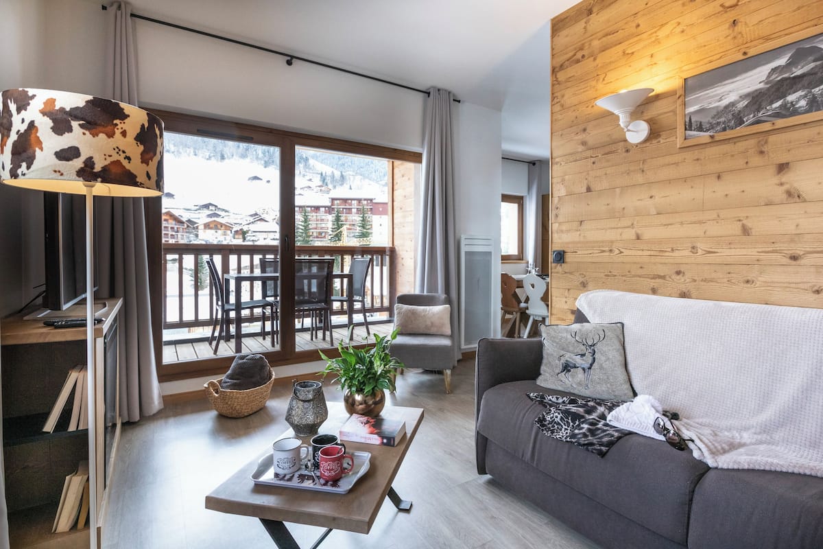 Een moderne woonkamer met een bank, een salontafel met een dienblad vol mokken, een lamp met een kousenvel lampscherm, en uitzicht op een besneeuwd berglandschap door grote ramen.