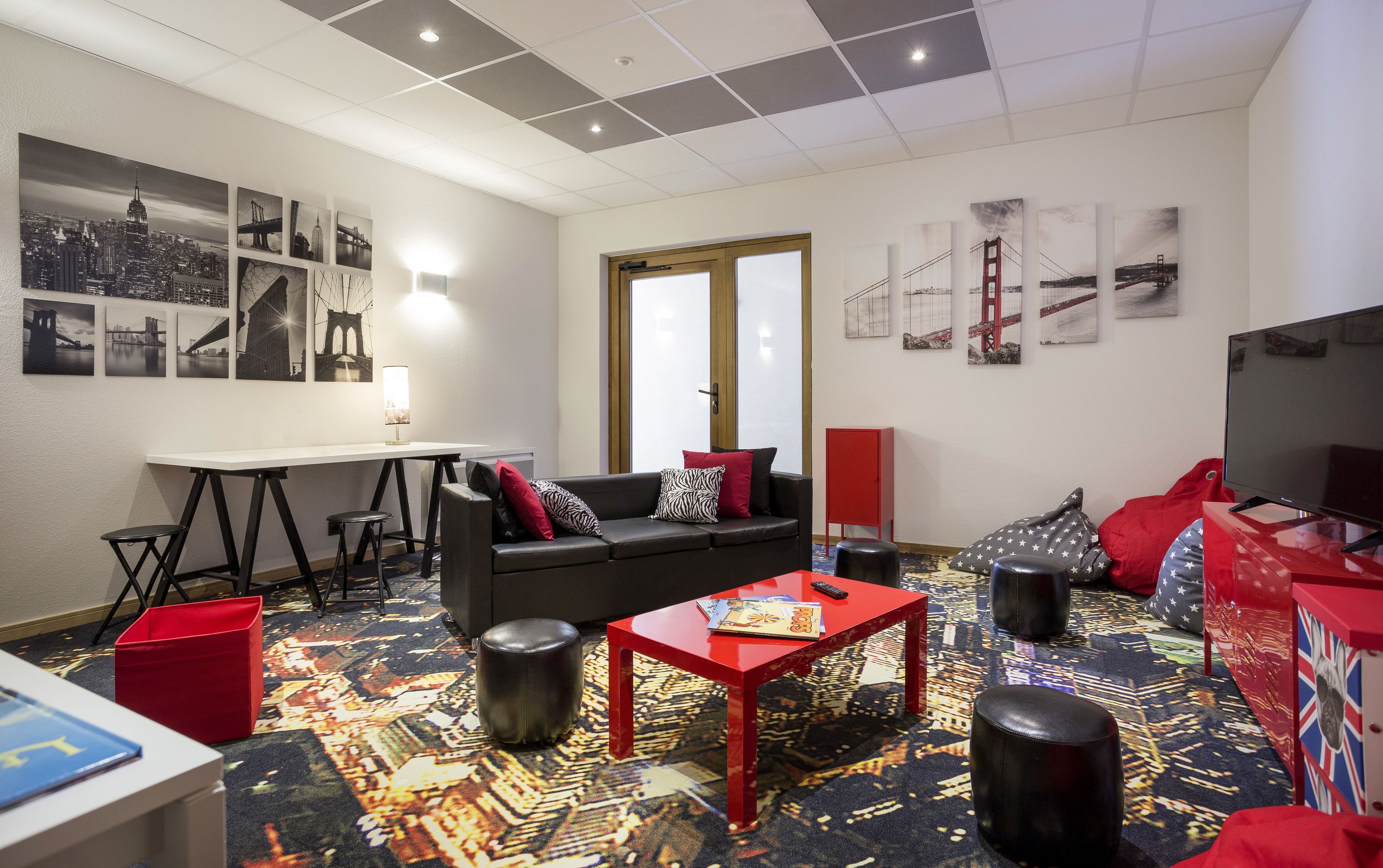 A modern hotel room with a black leather sofa, red accents, a flat-screen TV, and cityscape artwork on the walls.