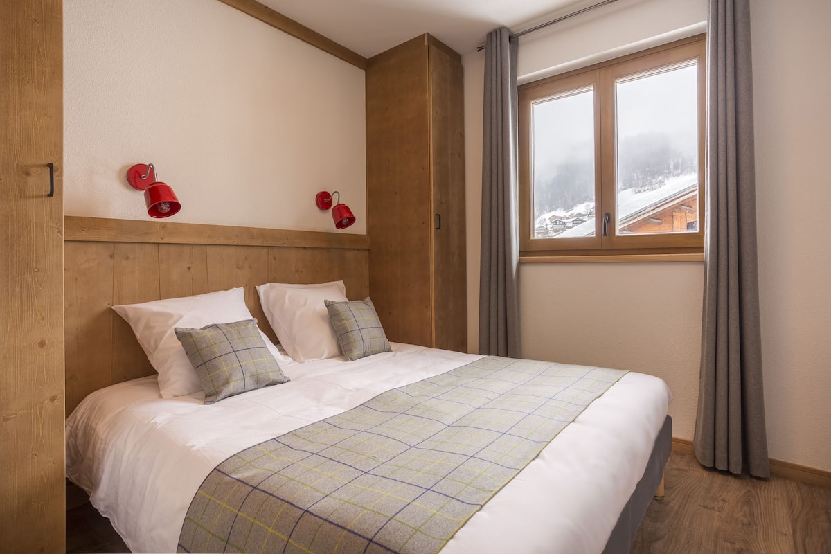 Een slaapkamer met een houten hoofdeinde, een bed met witte beddengoed en een geruite deken, twee rode wandlampen en een raam met gordijnen.