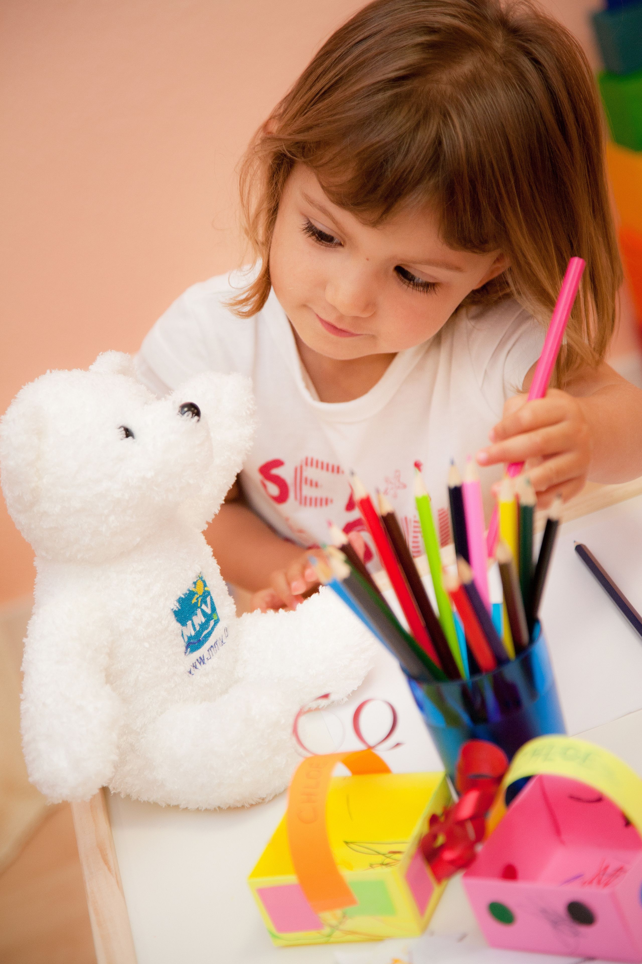 Ein Kind sitzt an einem Tisch mit einem weißen Teddybären und einer Auswahl bunter Stifte.