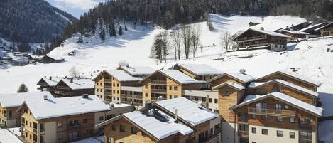 Un complejo turístico cubierto de nieve con varias edificaciones de madera, una pista de esquí al fondo y un cielo despejado.