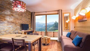 A room with a wooden table, chairs, a sofa, and a large window with a mountain view. - Résidence Club MMV l'Altaviva (Tignes)