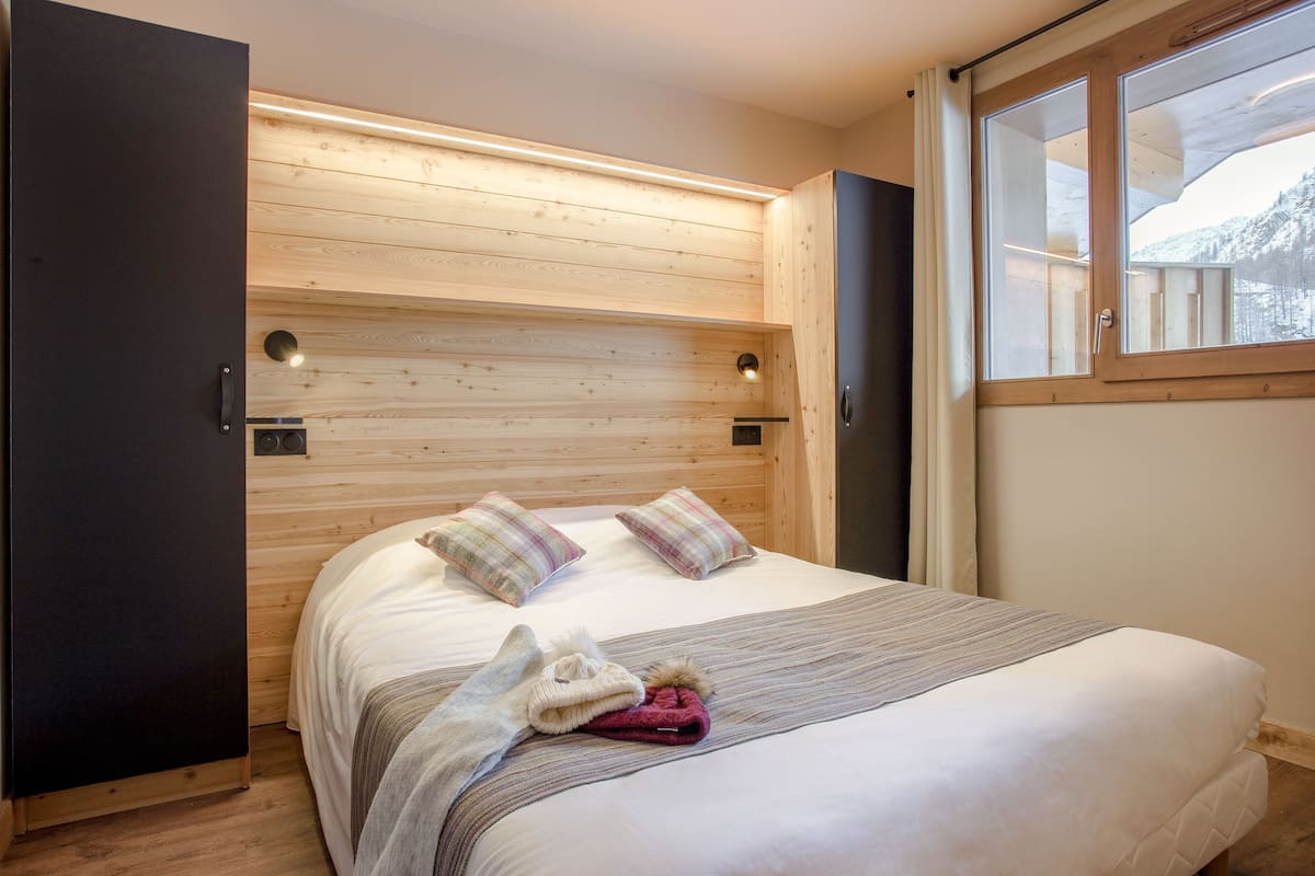 Un dormitorio con cabecera de madera, cama con dos almohadas, ventana con vistas a un paisaje nevado y una puerta a la izquierda.