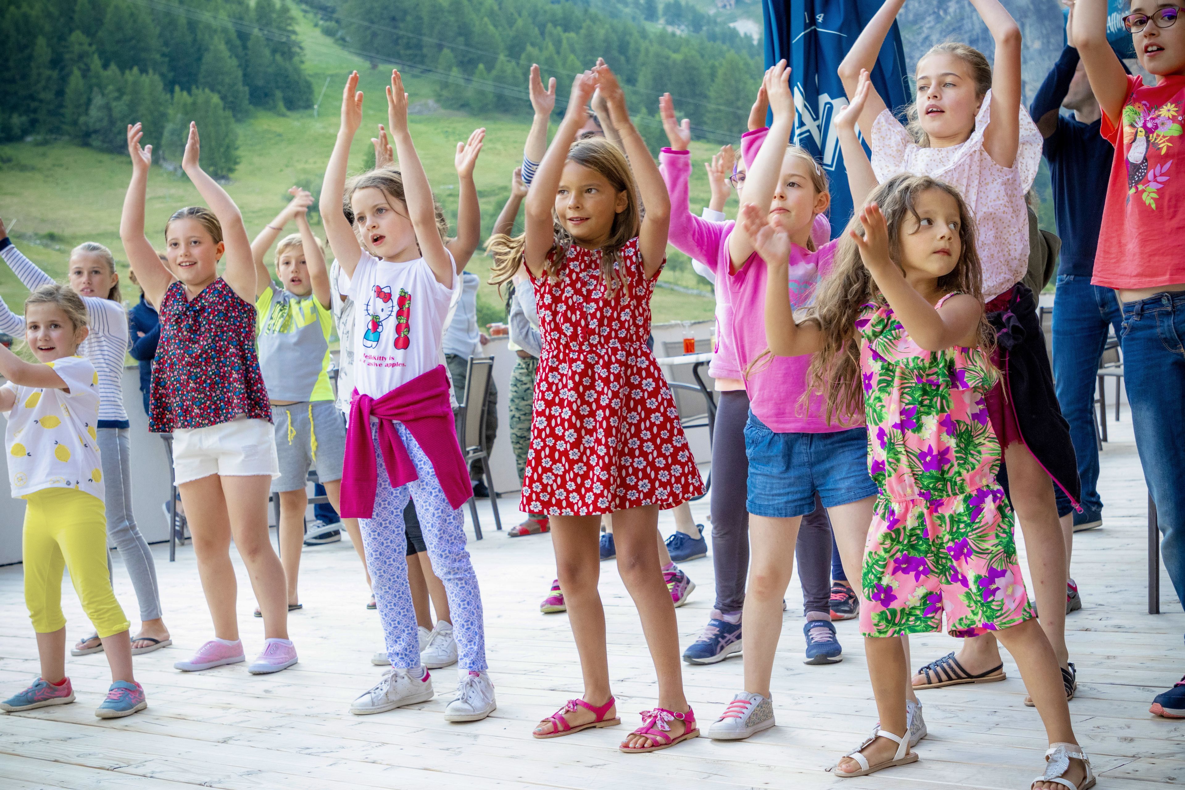 Eine Gruppe von Kindern nimmt draußen an einer Aktivität auf einer Holzterrasse teil, im Hintergrund ist eine grasbewachsene Hügelkuppe zu sehen.