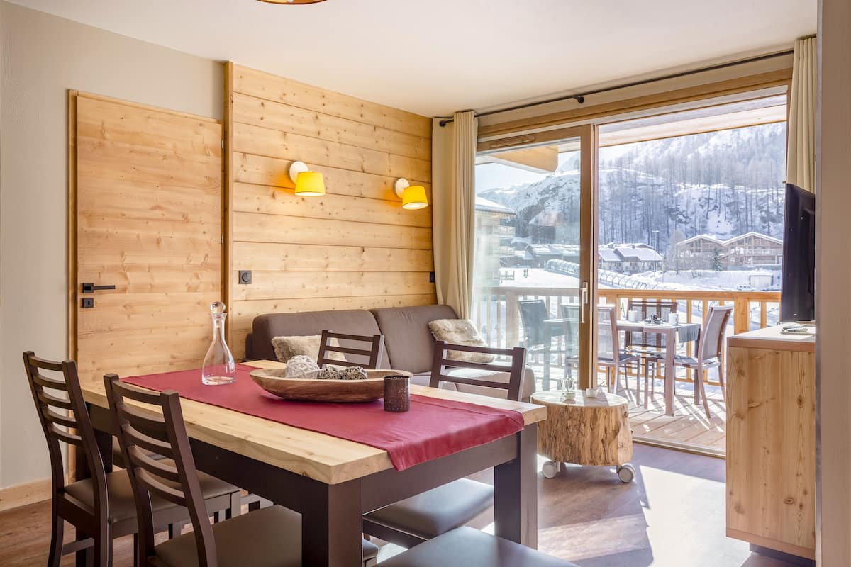 Un comedor con una mesa de madera, sillas y vistas a un paisaje nevado de montaña a través de grandes ventanales.