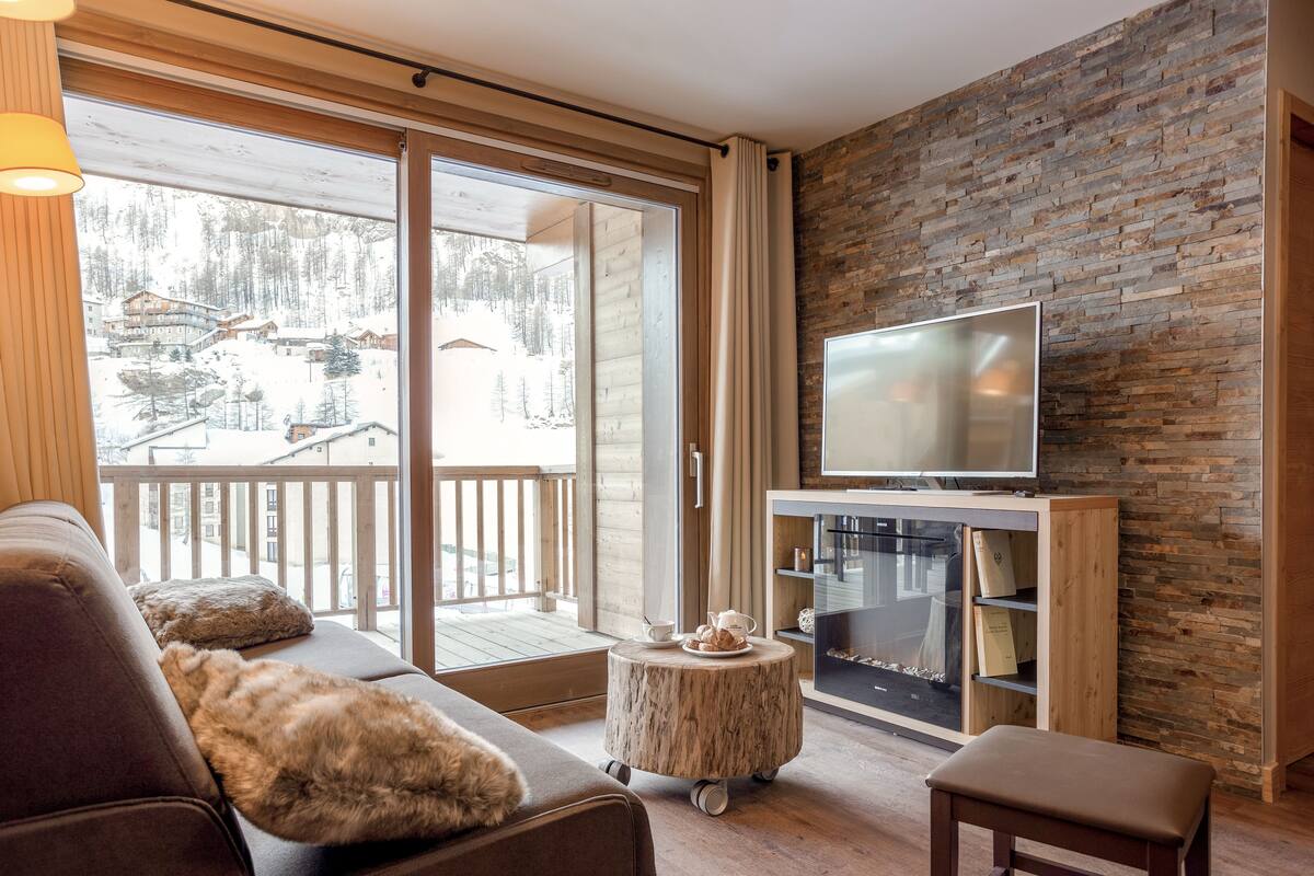 Una sala de estar acogedora con un televisor grande, muebles de madera y vistas a una zona montañosa nevada.