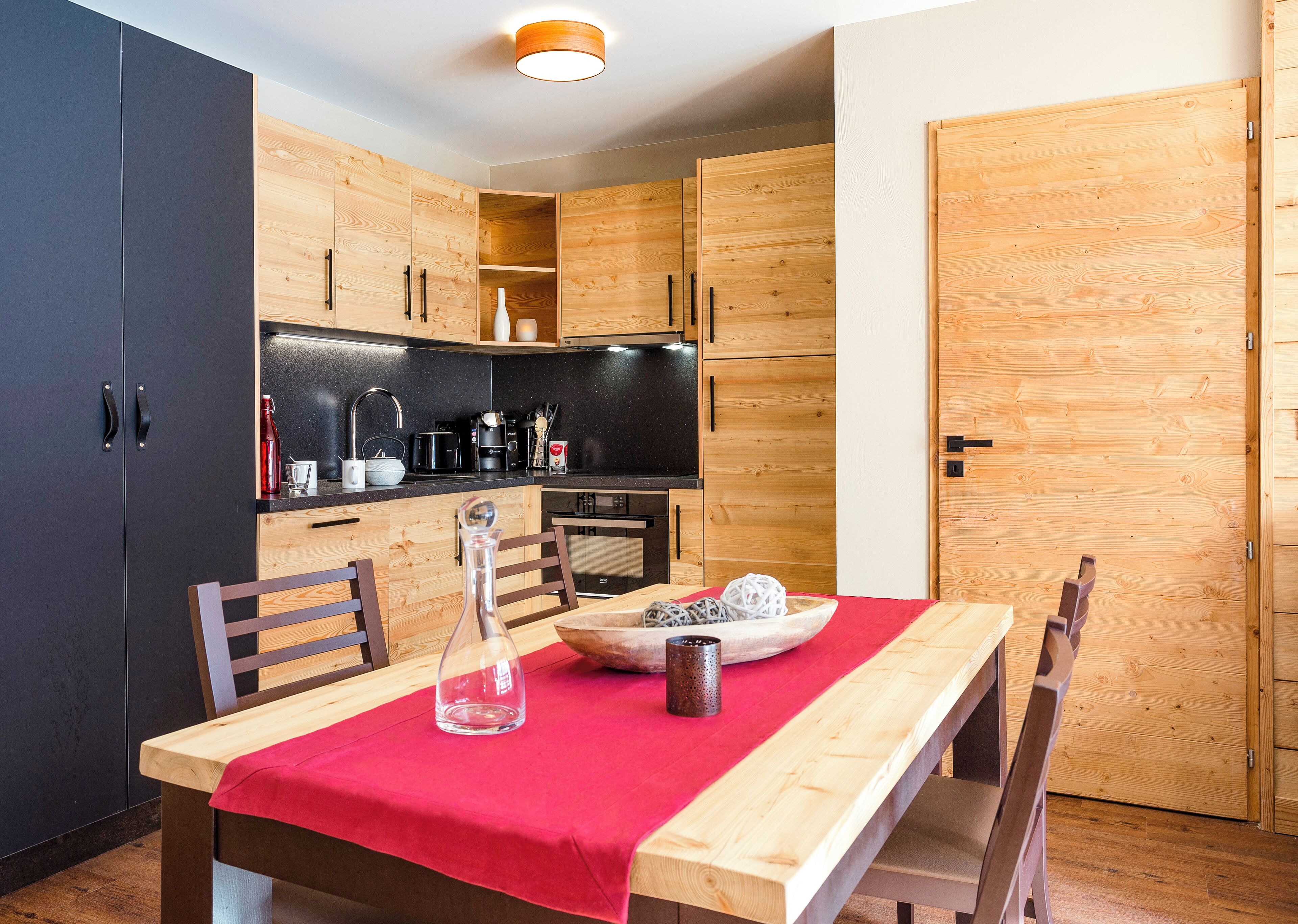 Una cucina compatta con mobili in legno, un piano di lavoro nero e un tavolo da pranzo con tovaglia rossa.