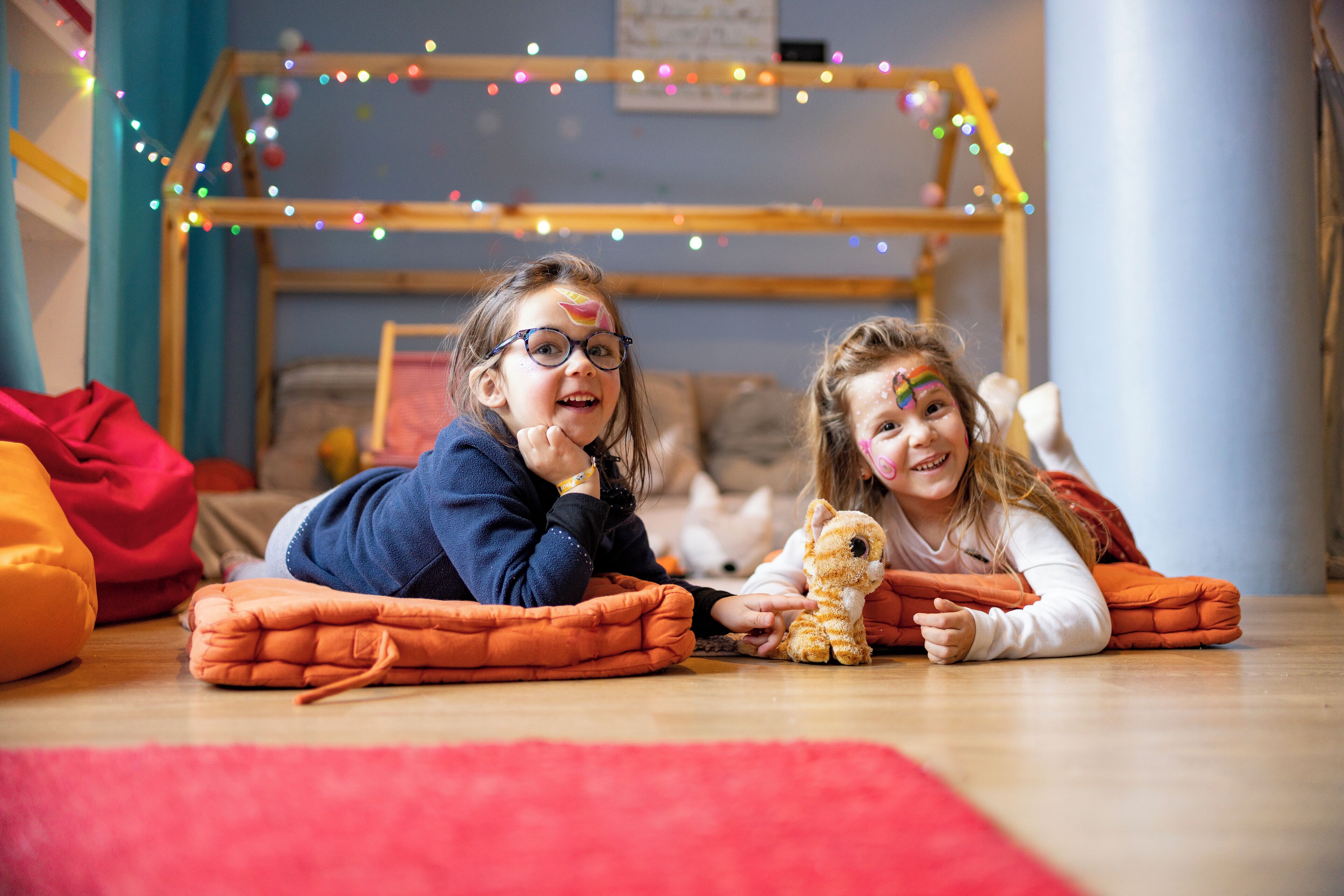 Two children lying on the floor with a stuffed toy, in a room with a wooden bed frame and string lights.