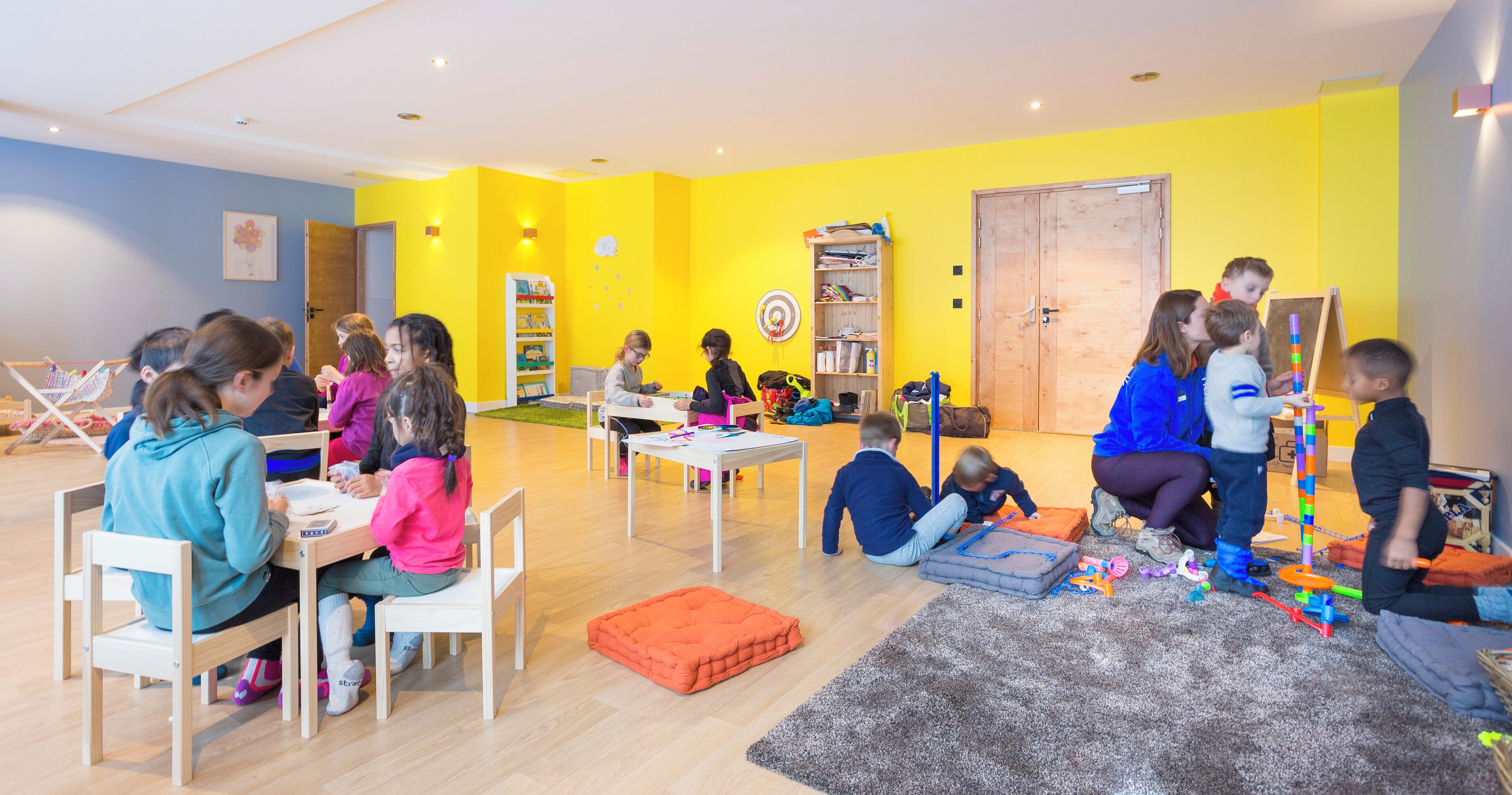 A brightly colored room with children engaged in various activities, including sitting at tables and playing on the floor.