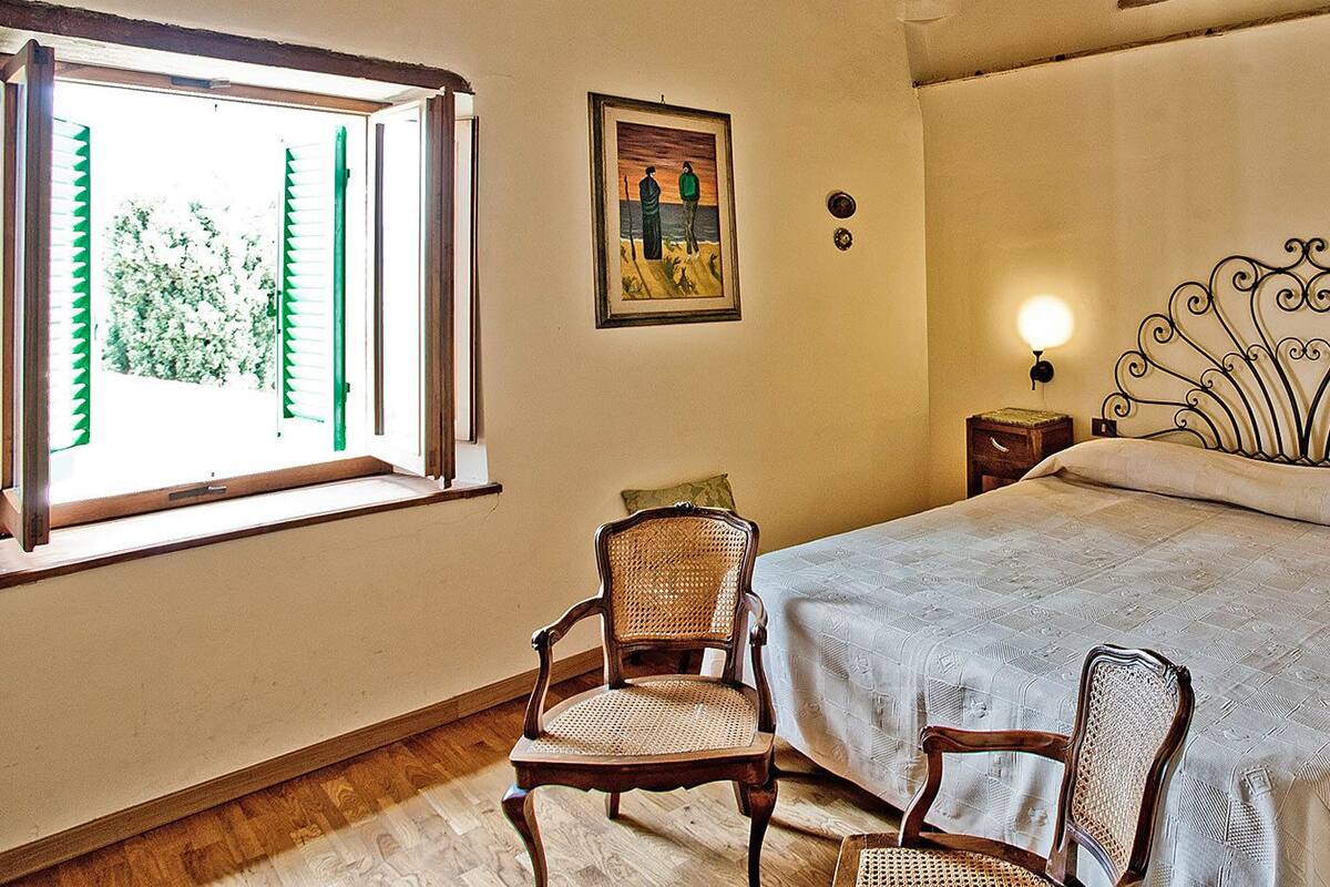 Ein Schlafzimmer mit einem Bett, zwei Nachttischen, einem Sessel, einem Fenster mit Jalousien und einem Wandbild.