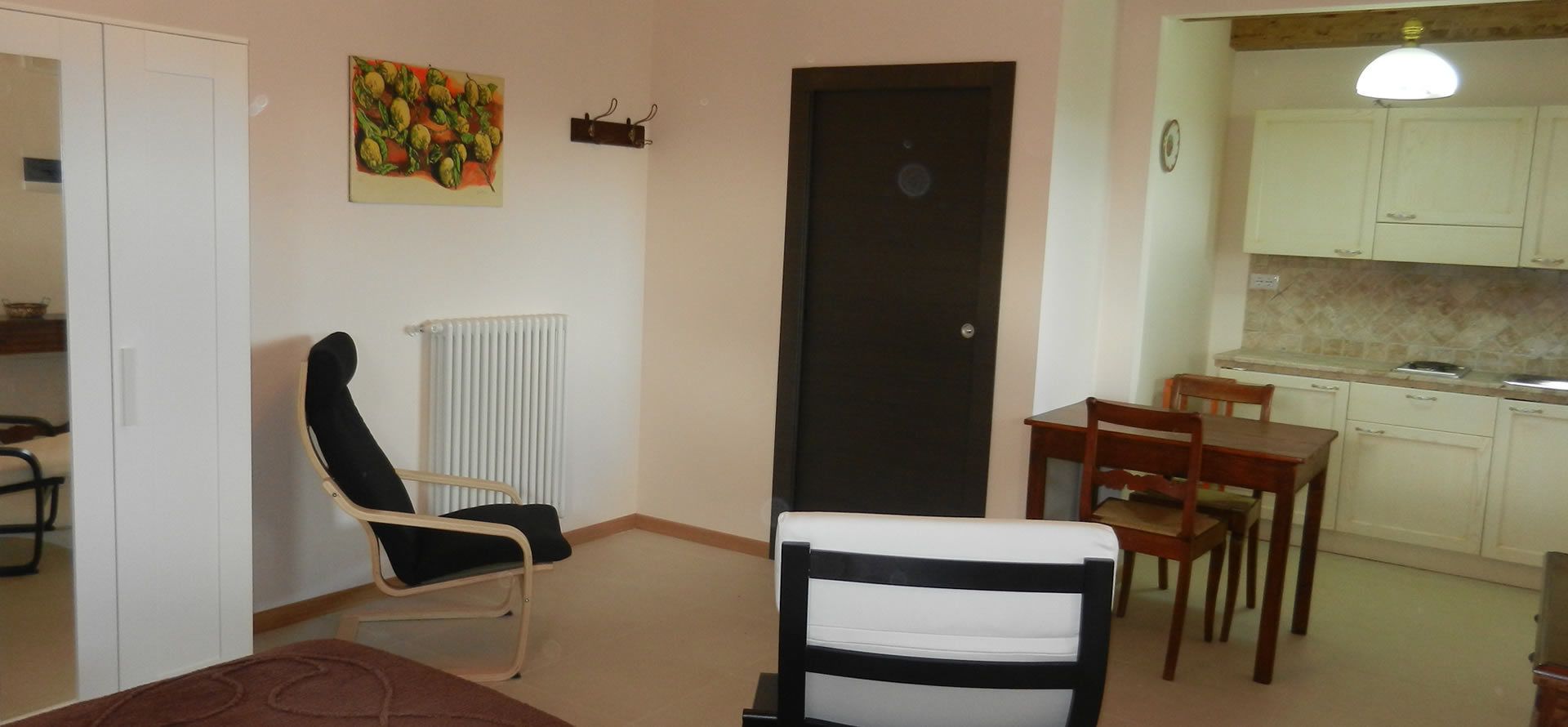 A room with a bed, a chair, a table, a radiator, a door, a kitchen area with cabinets and a countertop.