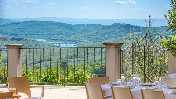 Un área de comedor al aire libre con mesas y sillas, vista a un lago y colinas, y una baranda de metal.