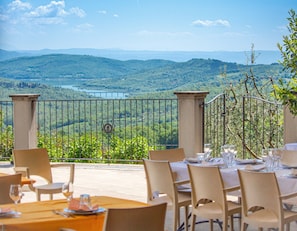 Un espace extérieur aménagé pour les repas, avec des tables et des chaises, une vue sur un lac et des collines, et une rambarde en métal.