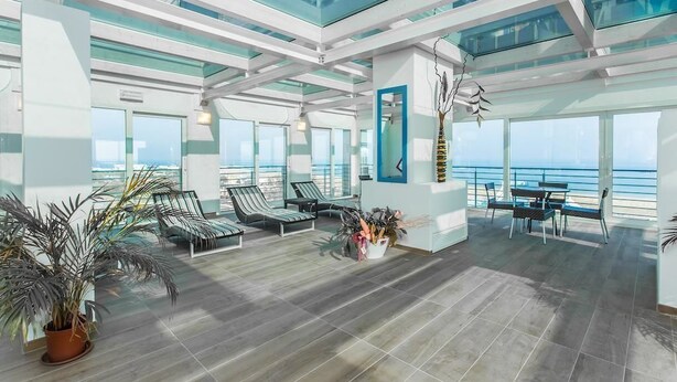 A spacious indoor area with a high ceiling, large windows, and a view of the ocean. There are several lounge chairs and a small table with chairs. - Lido Hotel Residence (Cesenatico)