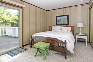 Un dormitorio con una cama de madera, un taburete verde, una mesita de noche, un cuadro y vistas al exterior con vegetación.
