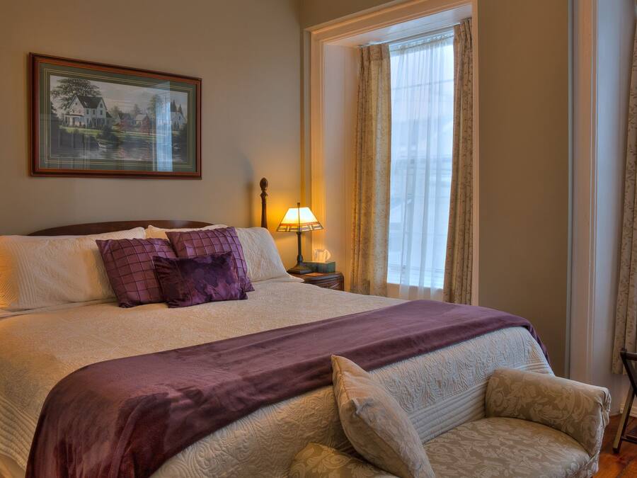 A neatly arranged bedroom with a large bed, a nightstand, a bedside table, a chair, and a window with curtains.