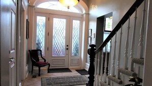 A hallway with a door, a chair, a rug, a staircase with a banister, and a chandelier. - Sir Isaac Brock B&B Luxury Suites (Brockville)