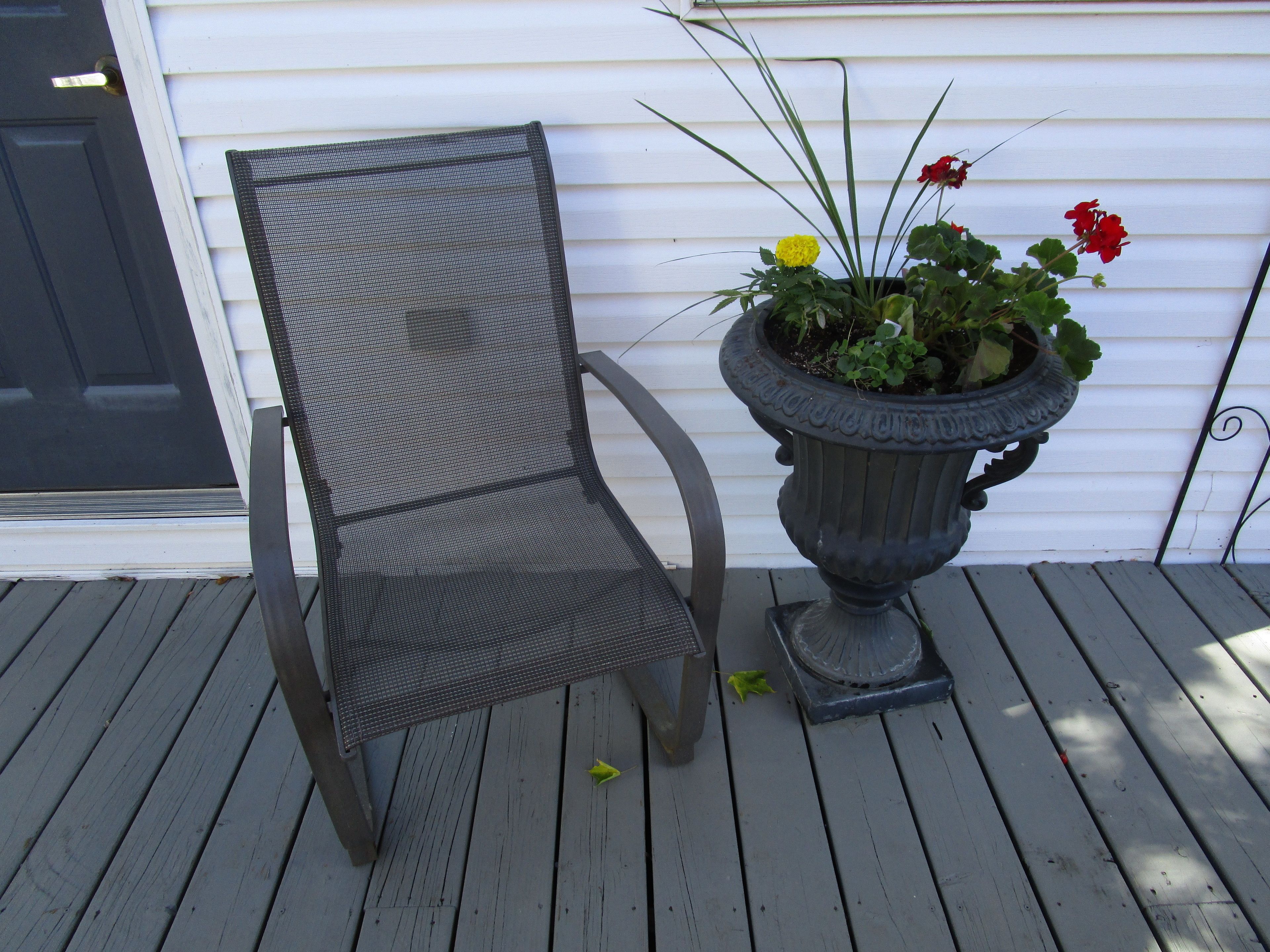 Une chaise en métal avec un dossier en treillis et une plante en pot aux fleurs rouges et jaunes, posée sur une terrasse en bois.