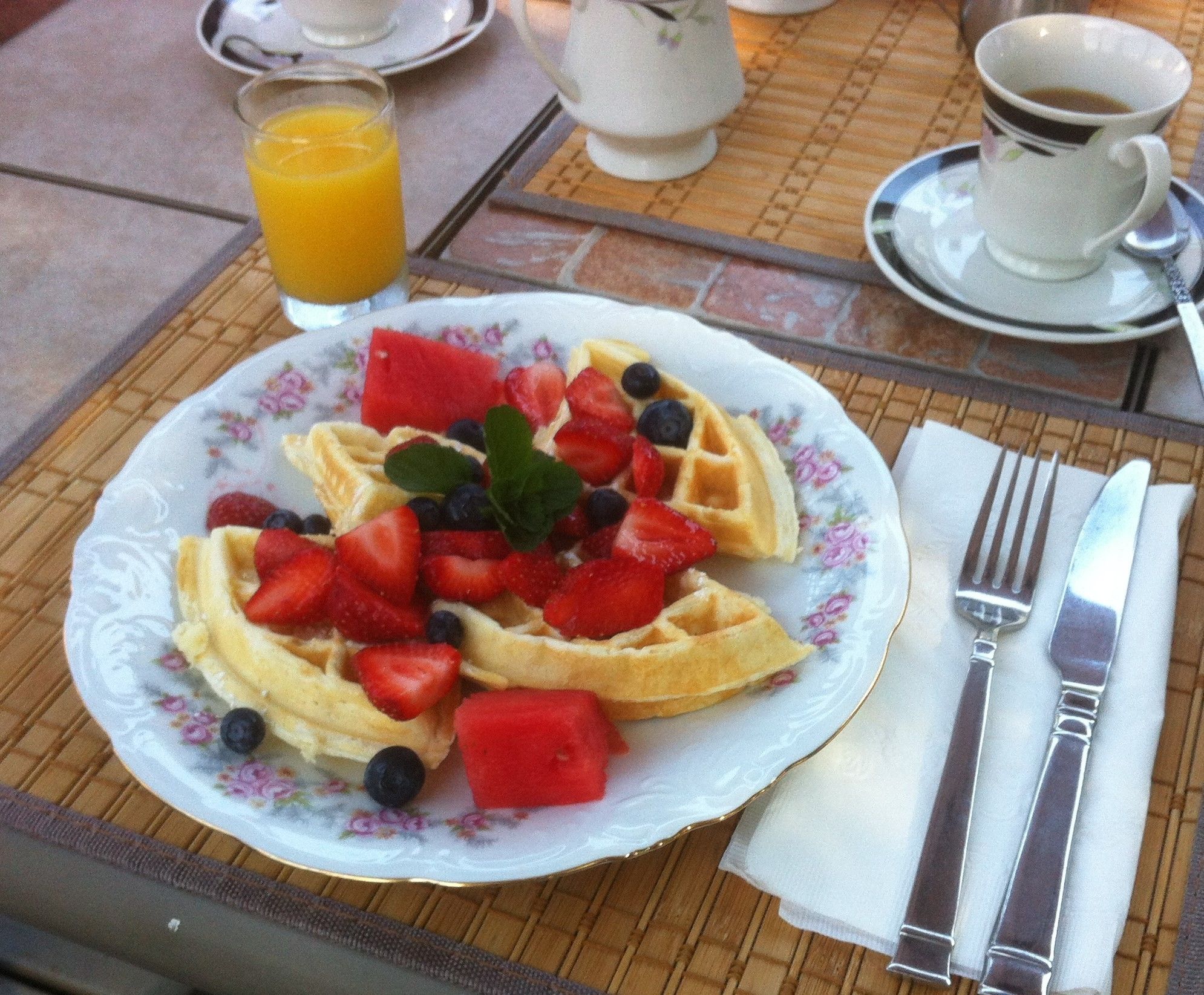 A plate of waffles topped with strawberries, blueberries, and watermelon, accompanied by a glass of orange juice and a cup of coffee on a table.
