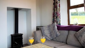 A living room with a sofa, two glasses of orange juice on a small table, a black wood-burning stove, and a window with floral curtains. - Kinbane Self-Catering (Ballycastle)