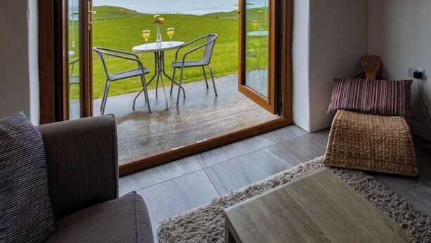 A room with a large sliding glass door leading to a grassy outdoor area with a view of the sea. Inside, there is a sofa, a small table with two glasses, and a television mounted on the wall. - Kinbane Self-Catering (Ballycastle)
