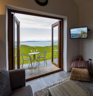 A room with a large sliding glass door leading to a grassy outdoor area with a view of the sea. Inside, there is a sofa, a small table with two glasses, and a television mounted on the wall. - Kinbane Self-Catering (Ballycastle)