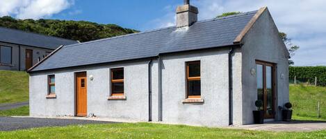 A small, single-story house with a grey exterior, a pitched roof, and a chimney. It has a wooden door and windows with wooden frames.