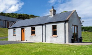A small, single-story house with a grey exterior, a pitched roof, and a chimney. It has a wooden door and windows with wooden frames. - Kinbane Self-Catering (Ballycastle)