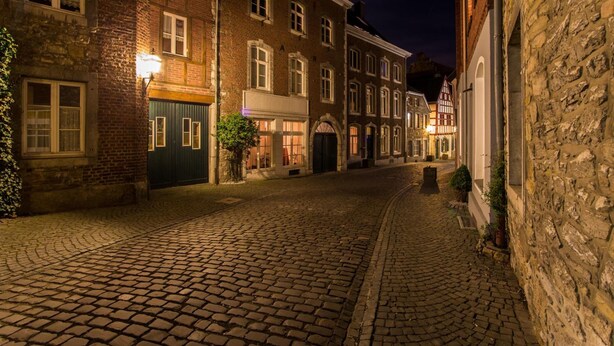 Uma rua de paralelepípedos ladeada por edifícios históricos, com fachadas de tijolo e pedra, e um estilo arquitetônico tradicional europeu.