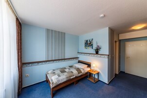 Un dormitorio con una cama individual, una mesita de noche con lámpara, una ventana con cortinas, una obra de arte colgada en la pared y una puerta.