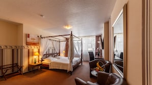 Uma cama com dossel e cortina, uma escrivaninha de madeira com abajur, uma poltrona de couro e um espaço de jantar com mesa e cadeiras.