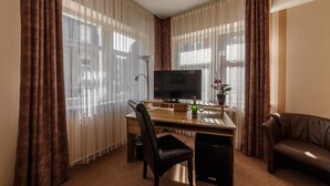 Quarto de hotel com escrivaninha de madeira, televisão, poltrona de couro preto, sofá de couro marrom e janelas amplas com cortinas translúcidas.