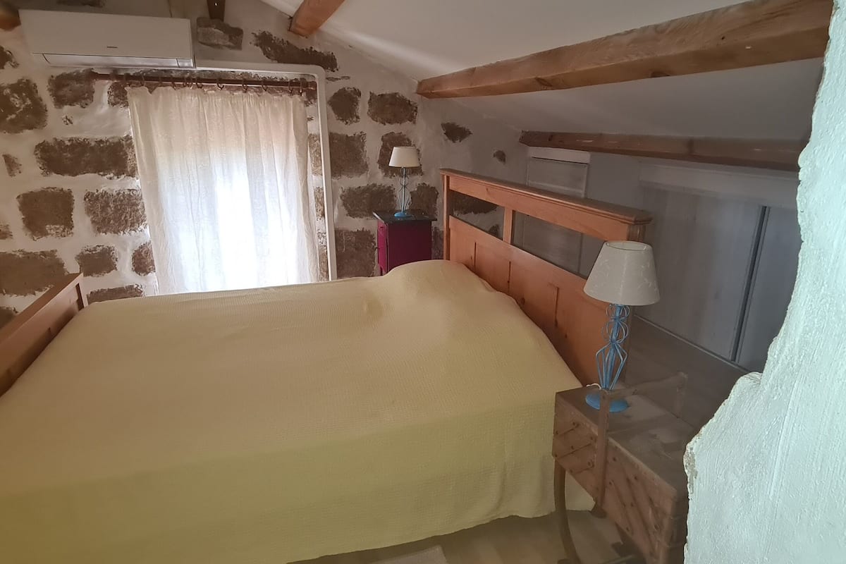 Un dormitorio con cama, una mesita de noche, una lámpara, una ventana con cortinas y una pared de piedra.