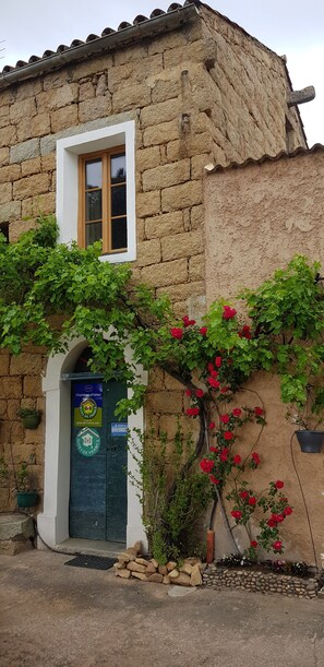 A stone building with a window and a door, surrounded by greenery and red flowers. - L'Orca Di San Gavinu (Figari)