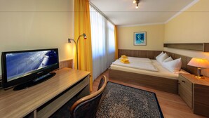 Una habitación de hotel con una cama, un escritorio con televisor, una silla, una mesita de noche con lámpara y una ventana con cortinas.